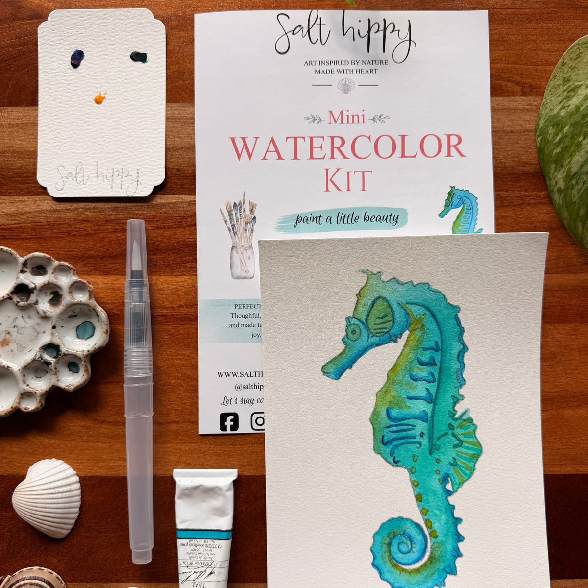 A mini watercolor kit by Salt Hippy displayed on a wooden surface with art supplies. The kit includes a watercolor paper card featuring a painted seahorse in teal and green, a tube of watercolor paper, a clear water brush.