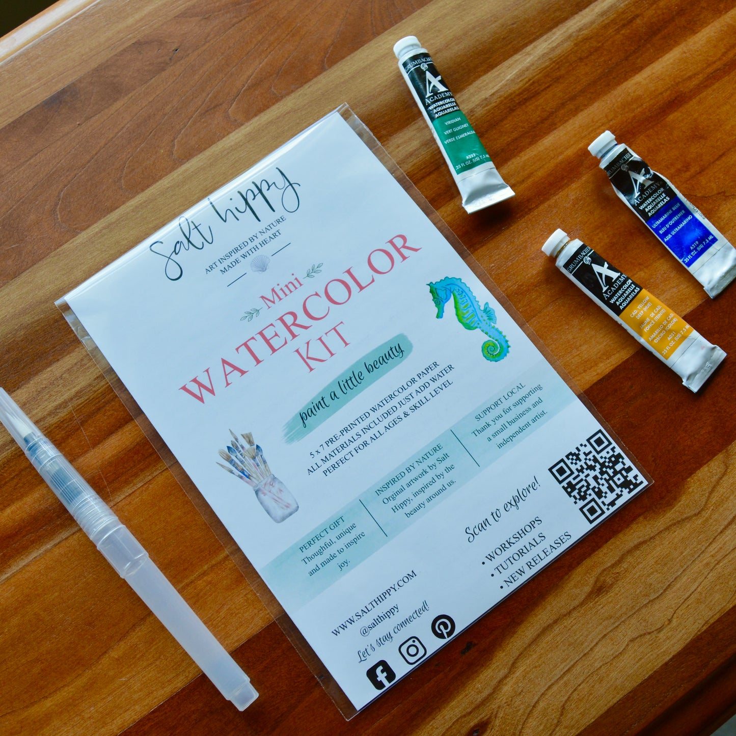 A 'Mini Watercolor Kit' by Salt Hippy, presented in a clear plastic sleeve on a wooden table, accompanied by a clear plastic paintbrush and three watercolor paint tubes.