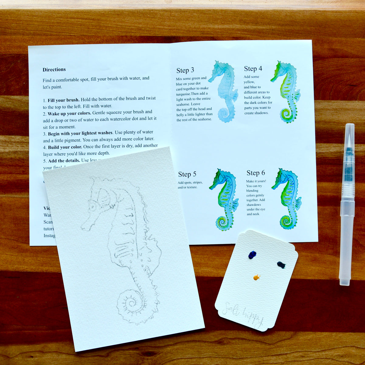 A seahorse-themed art kit laid out on a wooden table, including printed instruction sheets with step-by-step painting directions, a white paintbrush, a small palette card with paint wells, and a white card with the word 'Salt Hippy' written on it.