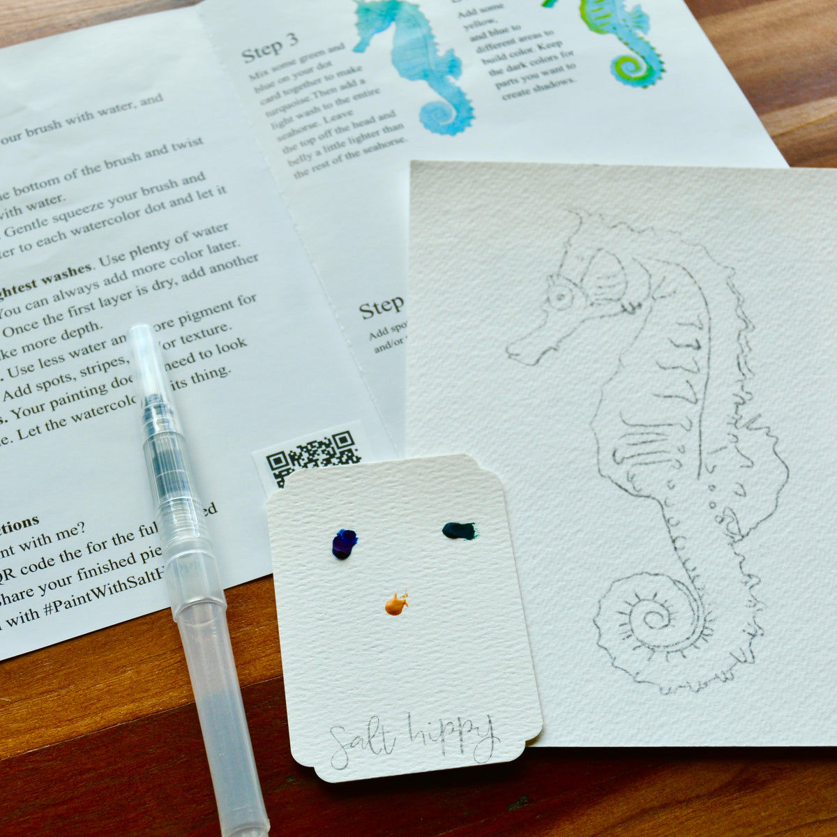 A close-up of a painting or printing setup on a wooden surface, showing printed instructions, a clear-handled brush, a small swatch card with paint dots, and a white sketch of a seahorse on textured paper.