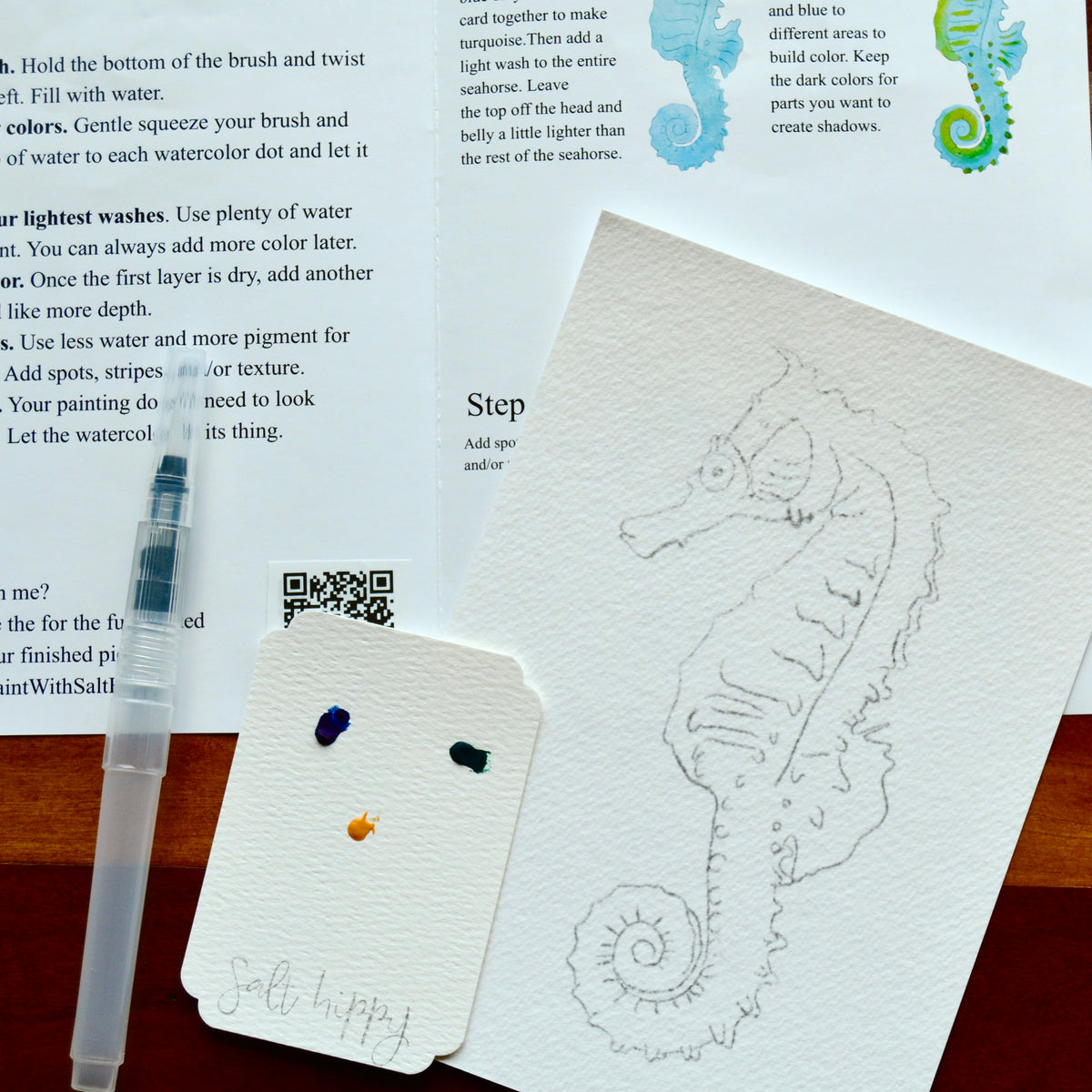 A small round white paint palette with wells holding blue, green, and yellow paint, placed on top of printed watercolor instruction sheets featuring seahorse illustrations.