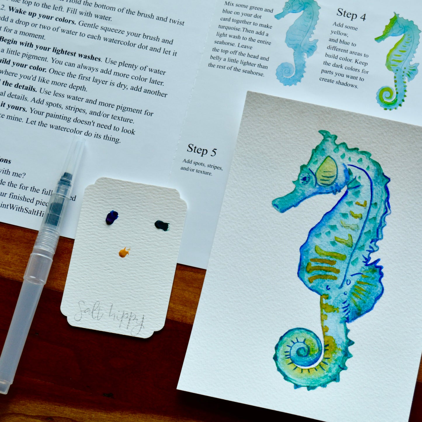 A close-up of a watercolor painting of a seahorse on textured paper, with printed painting instructions visible in the background. A small palette with paint tubes and a wooden palette stand sits on a tabletop in the lower left.