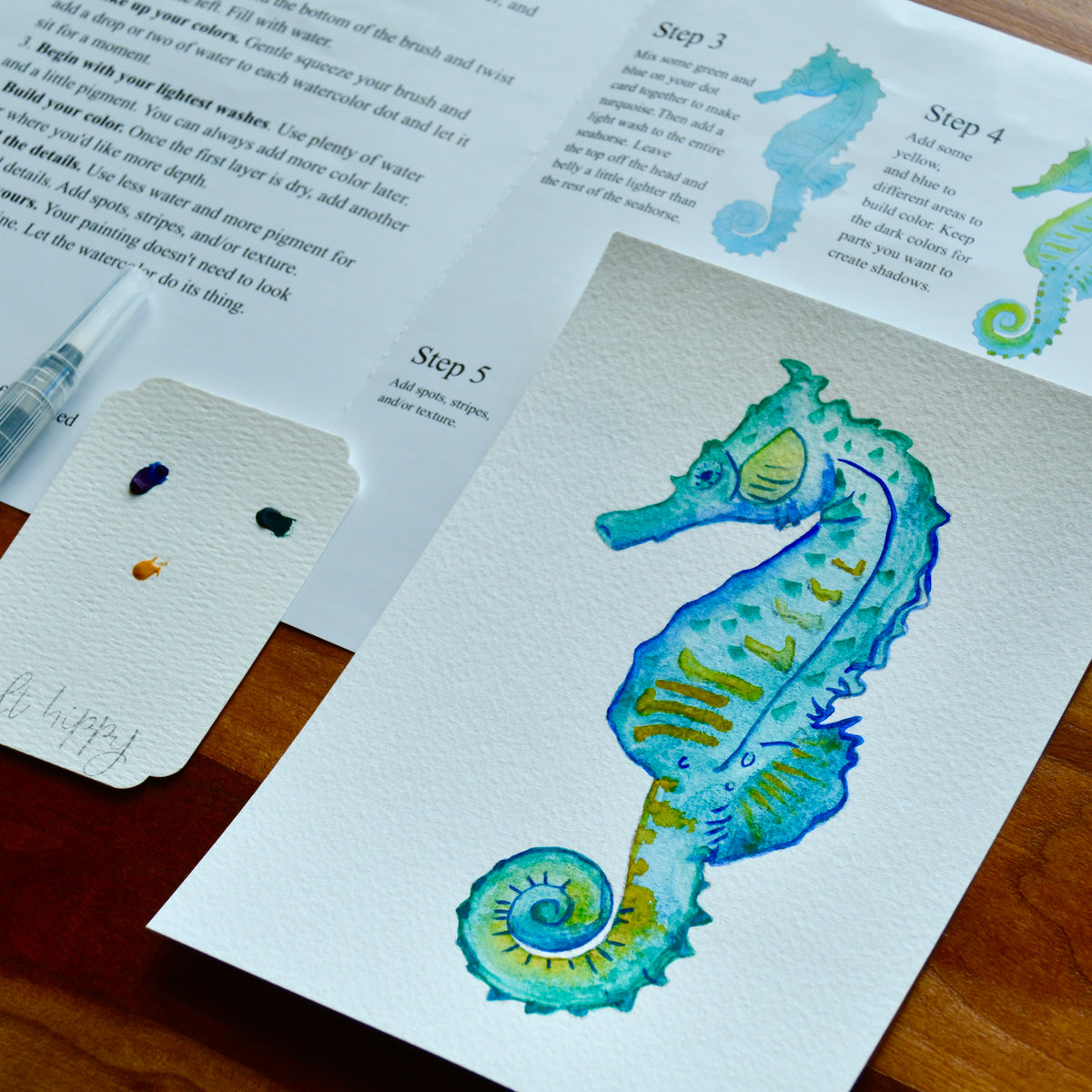 A watercolor painting of a seahorse on textured paper, shown alongside an instruction booklet with steps and a paint palette on a wooden surface.