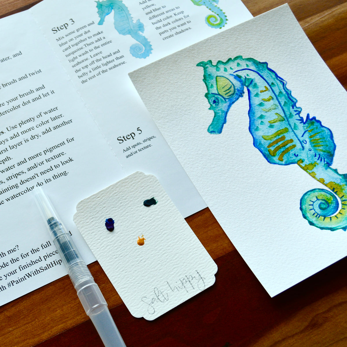 A watercolor seahorse art print displayed on a wooden table next to its printed making instructions, a small swatch card with paint colors, and a water brush.