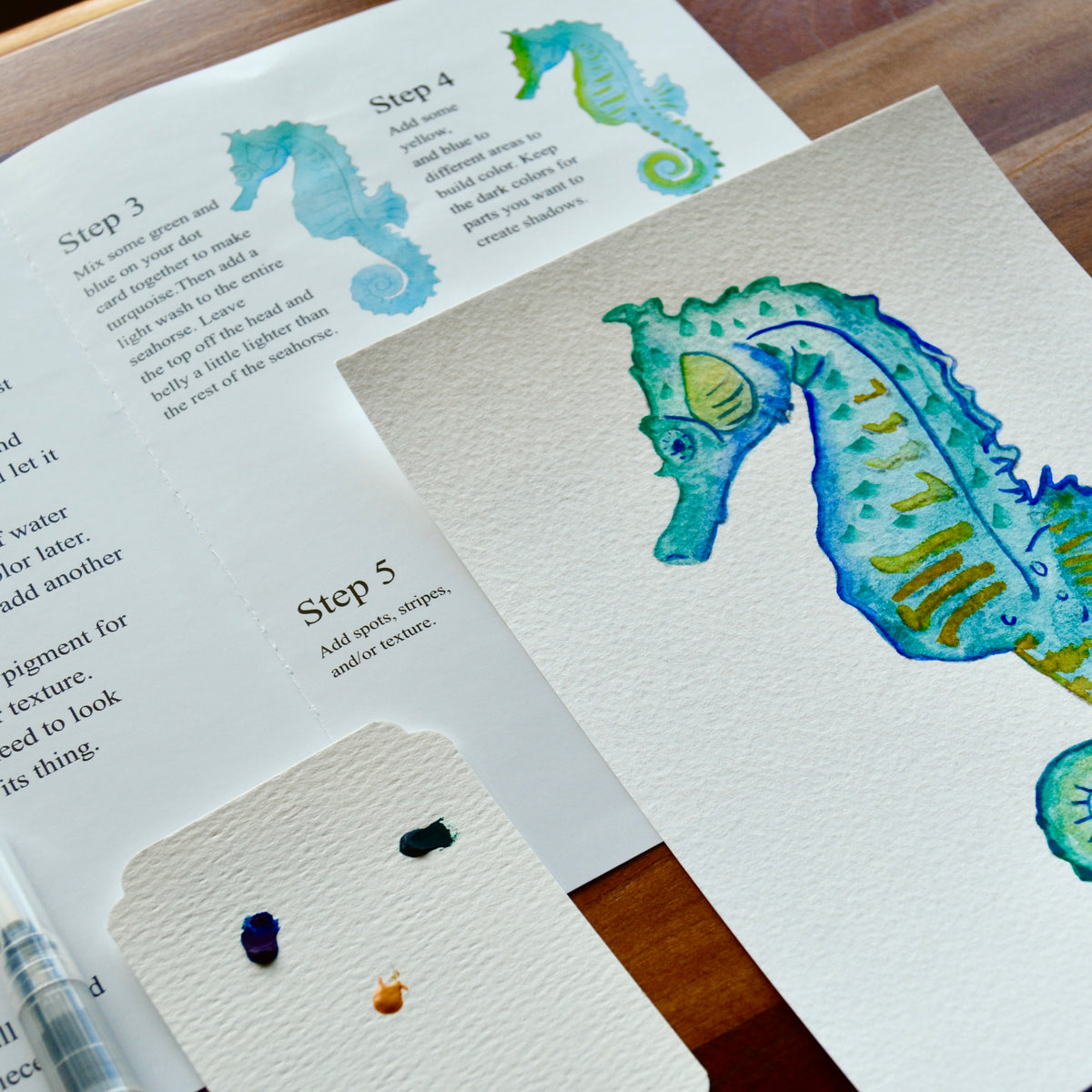 A watercolor seahorse artwork on textured white paper, shown alongside an open instruction sheet with step-by-step painting directions and paint swatches on a wooden surface.