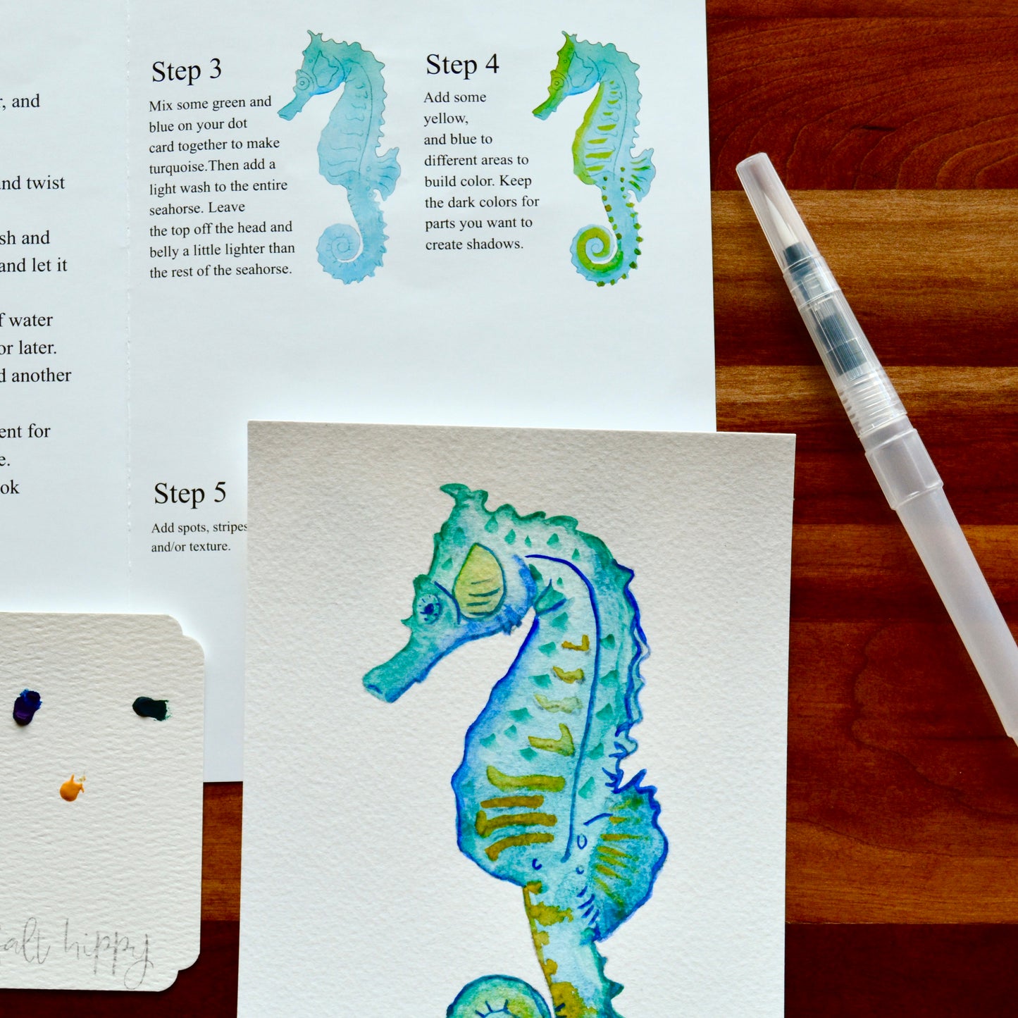 A watercolor painting of a seahorse on textured paper, shown alongside an instruction sheet with steps, a paint brush, and a wooden table background.