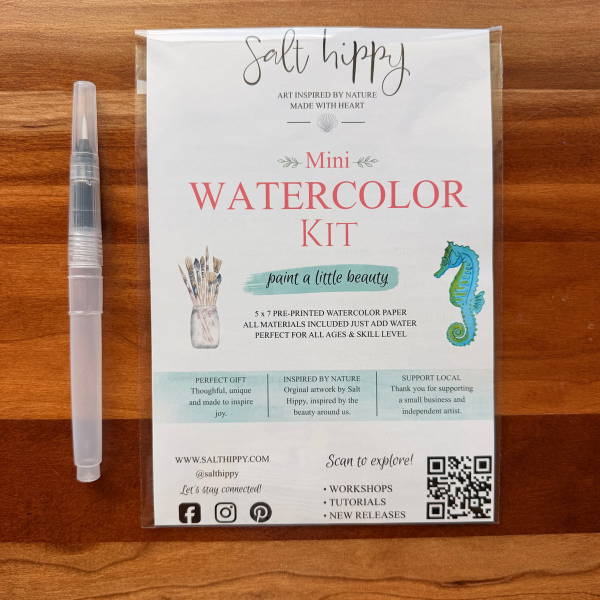 A Salt Hippy Mini Watercolor Kit in a clear plastic sleeve, displayed on a wooden surface next to a clear water brush. The packaging features a white background with pink and teal text, along with illustrations of a paintbrush and a seahorse.