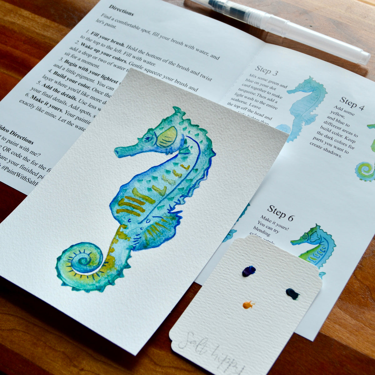 A close-up of a watercolor painting of a seahorse on textured paper, with printed instructions and swatches laid out on a wooden table.