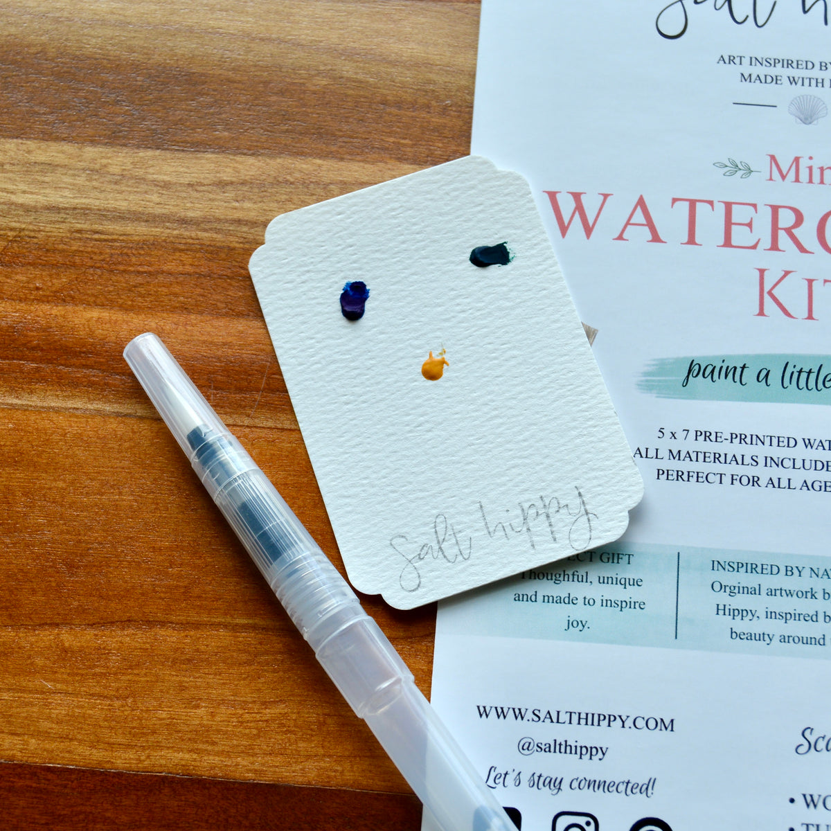 A clear water brush pen with blue ink lies on a wooden tabletop next to a small white card showing three paint colors, with printed paper underneath.